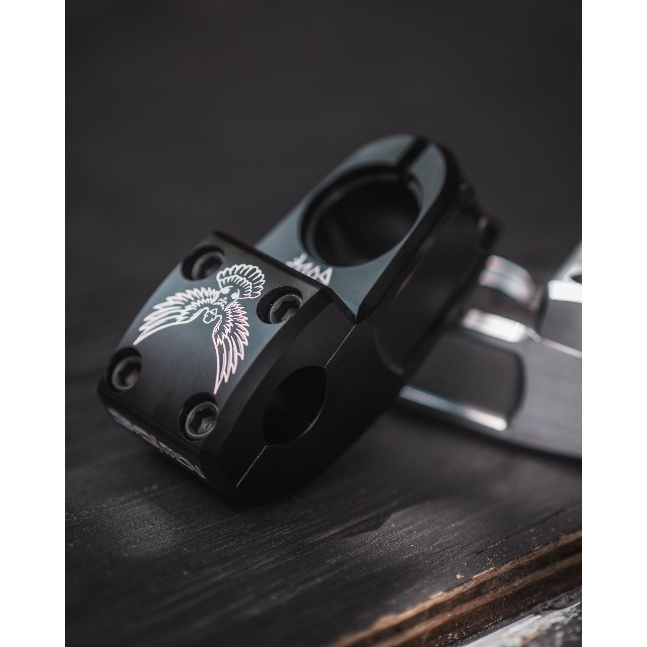 A close-up of the black metal Demolition Josh Dove Stem, featuring a white winged emblem design, rests elegantly on a dark wooden surface.