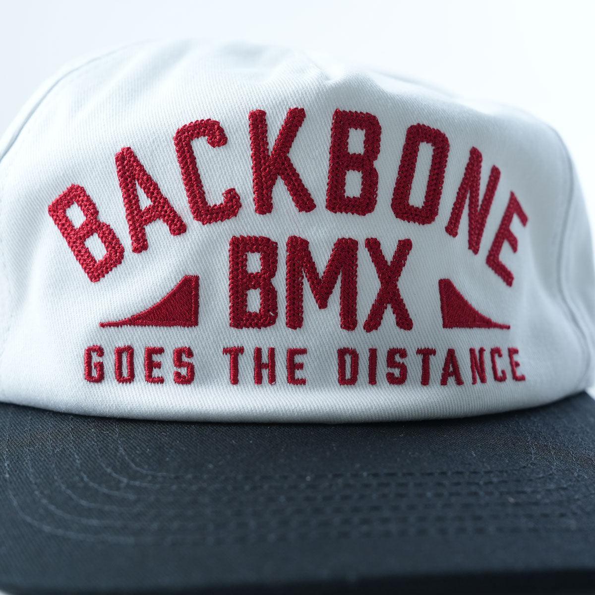 The Backbone Distance Snapback Hat is a white and black snapback featuring red embroidered text that reads “BACKBONE BMX GOES THE DISTANCE.”.