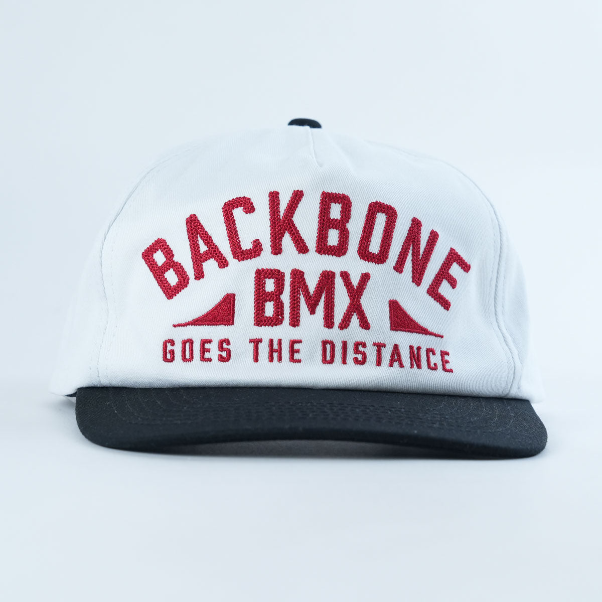 The Backbone Distance Snapback Hat is a white, unstructured 5-panel cap with a black brim, featuring red embroidered text that reads “BACKBONE BMX GOES THE DISTANCE.”.