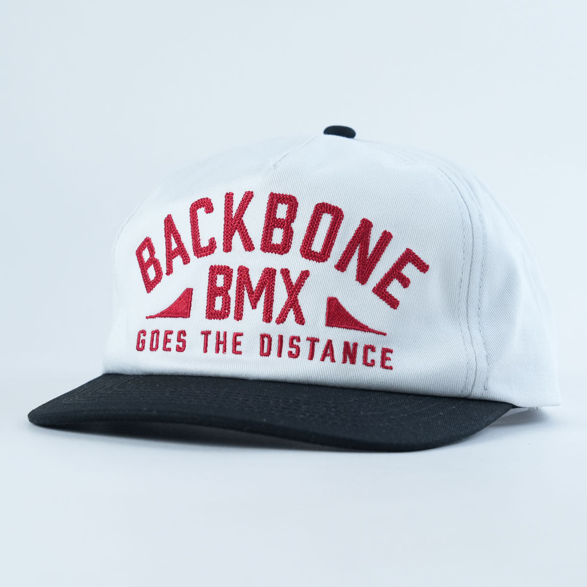 The Backbone Distance Snapback Hat is an unstructured white and black cap featuring “BACKBONE BMX GOES THE DISTANCE” in red letters on the front.