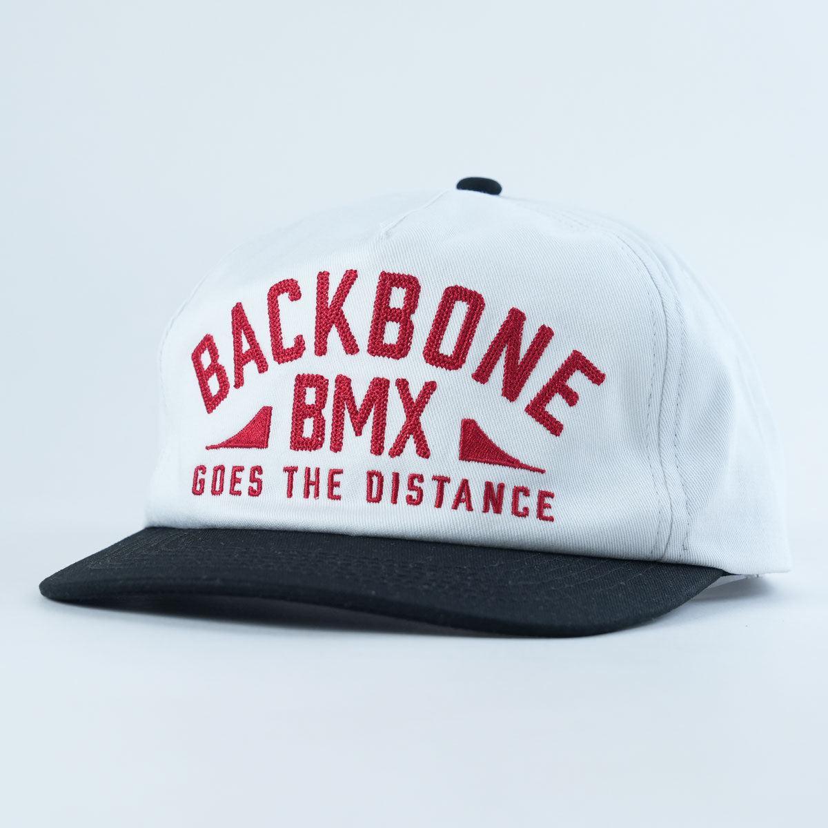 The Backbone Distance Snapback Hat is an unstructured white and black cap featuring “BACKBONE BMX GOES THE DISTANCE” in red letters on the front.