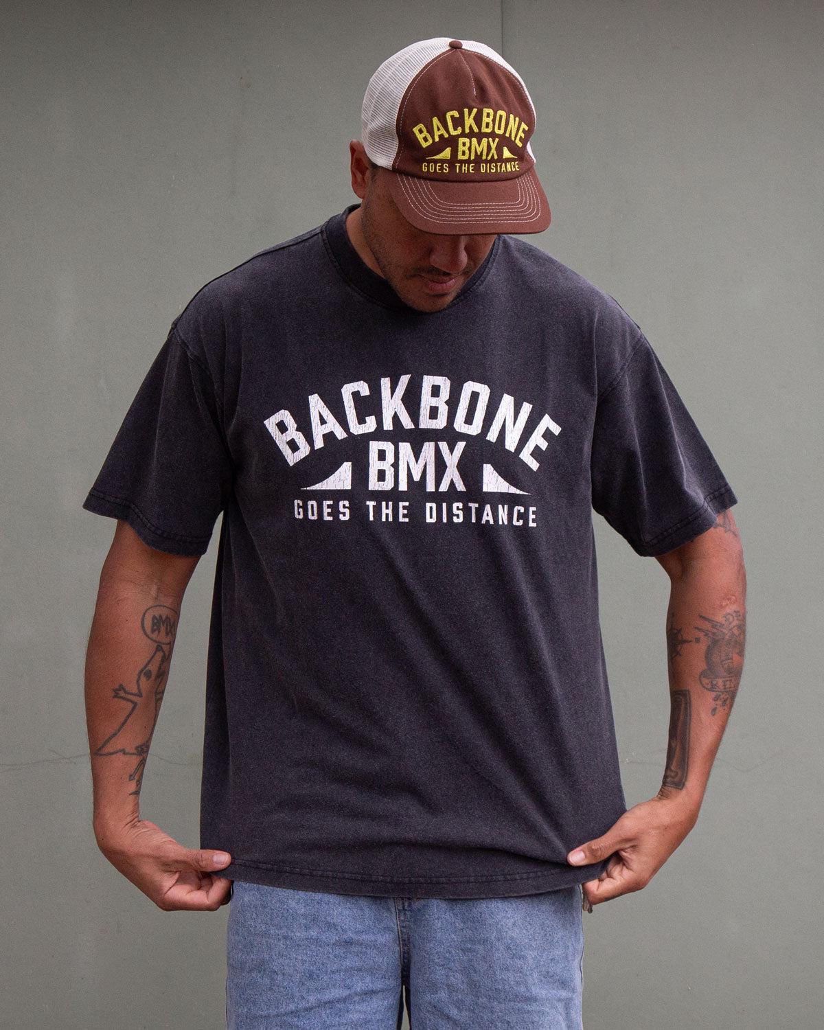 A man wearing the Backbone Distance Vintage T-Shirt and a cap stands against a plain background, looking down while holding the bottom of his shirt.