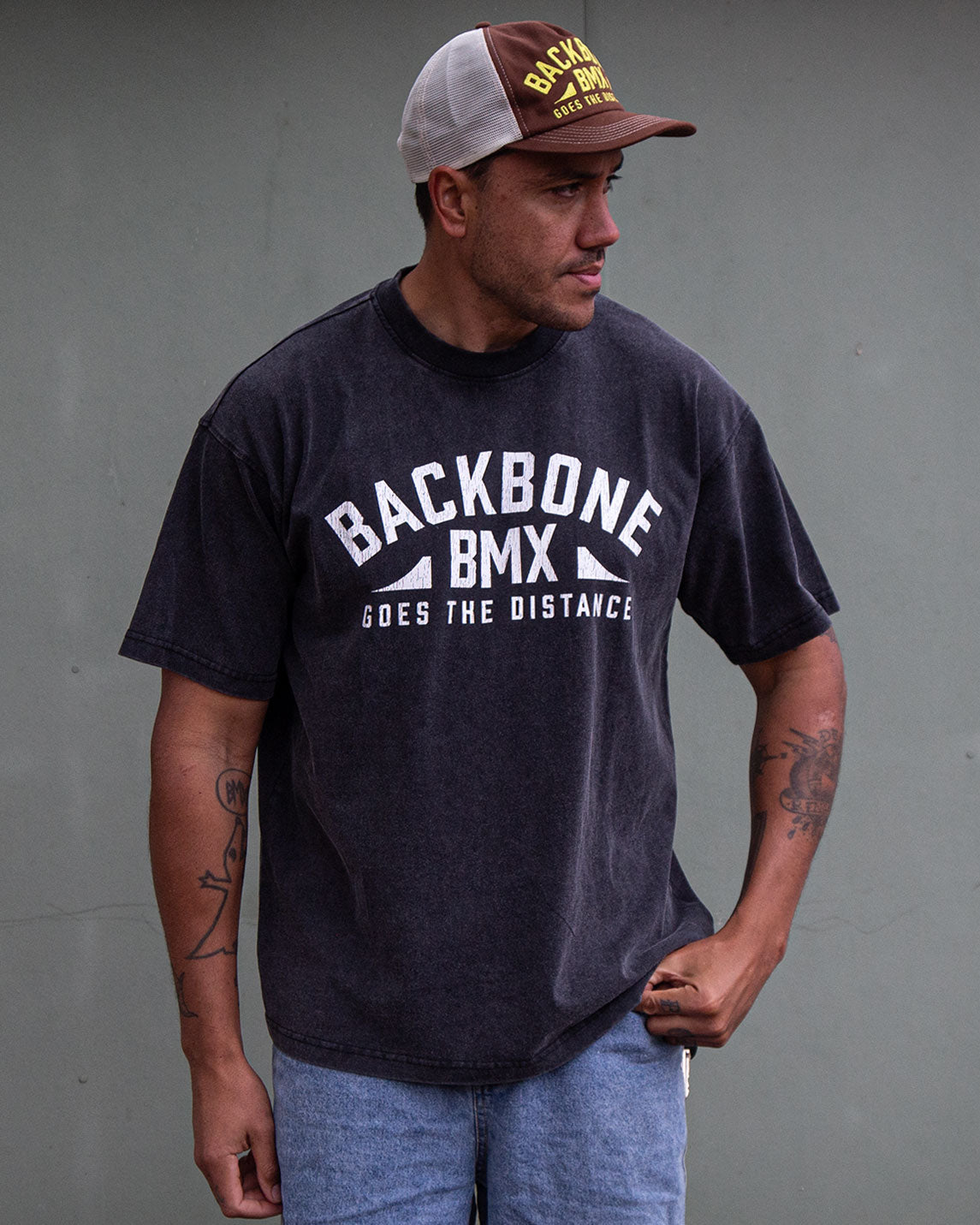 A man in a premium cotton Backbone Distance Vintage T-Shirt and a trucker hat stands against a plain grey wall, looking to his left.