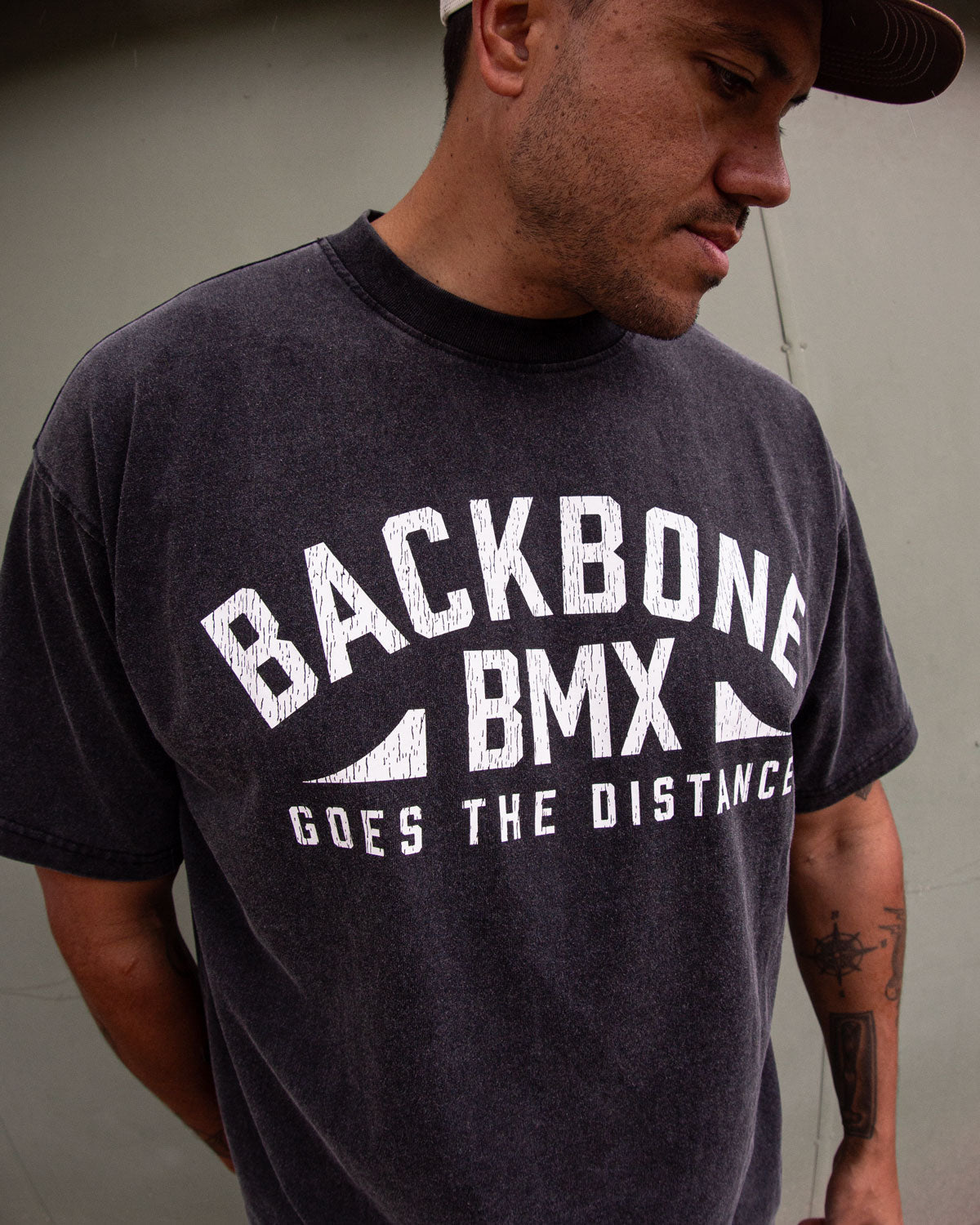 Wearing the Backbone Distance Vintage T-Shirt featuring the text “BACKBONE BMX GOES THE DISTANCE,” a person stands against a plain wall, gazing down and to the side.