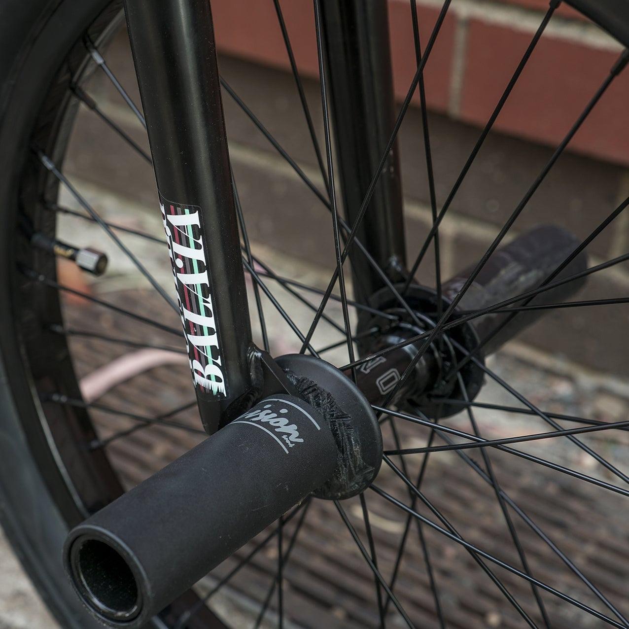 Close-up of a black bicycle wheel with metal spokes and a nearby Division Dynamic Plastic Peg (Each) / 10-14mm Black on an outdoor pavement, with a brick wall in the background.