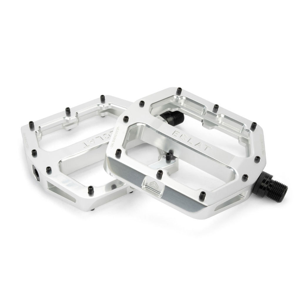 Two silver Eclat Surge XL Pedals with black bolts feature a flat design and multiple grip pins. The oversize platform area ensures stability, while the left pedal is slightly tilted, showing both the top and side views.
