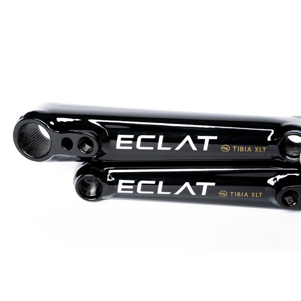 Close-up of two black metal components with the brand name "ECLAT" and model "Eclat Tibia XLT Cranks" printed on them. The lightweight crank parts have holes on both ends and reflective surfaces, making them perfect for a BMX crankset.