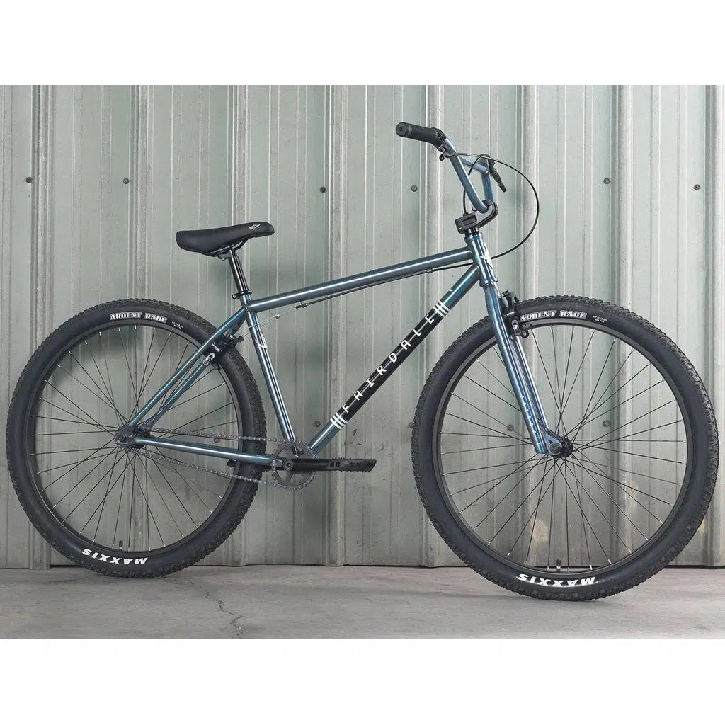 A Fairdale Taj 27.5 Inch Bike (2022) with large blue tires stands against a corrugated metal wall. The chromoly frame features the brand "Throne," and the tires are branded "Maxxis." This bike comes equipped with a single-speed drivetrain for streamlined performance.