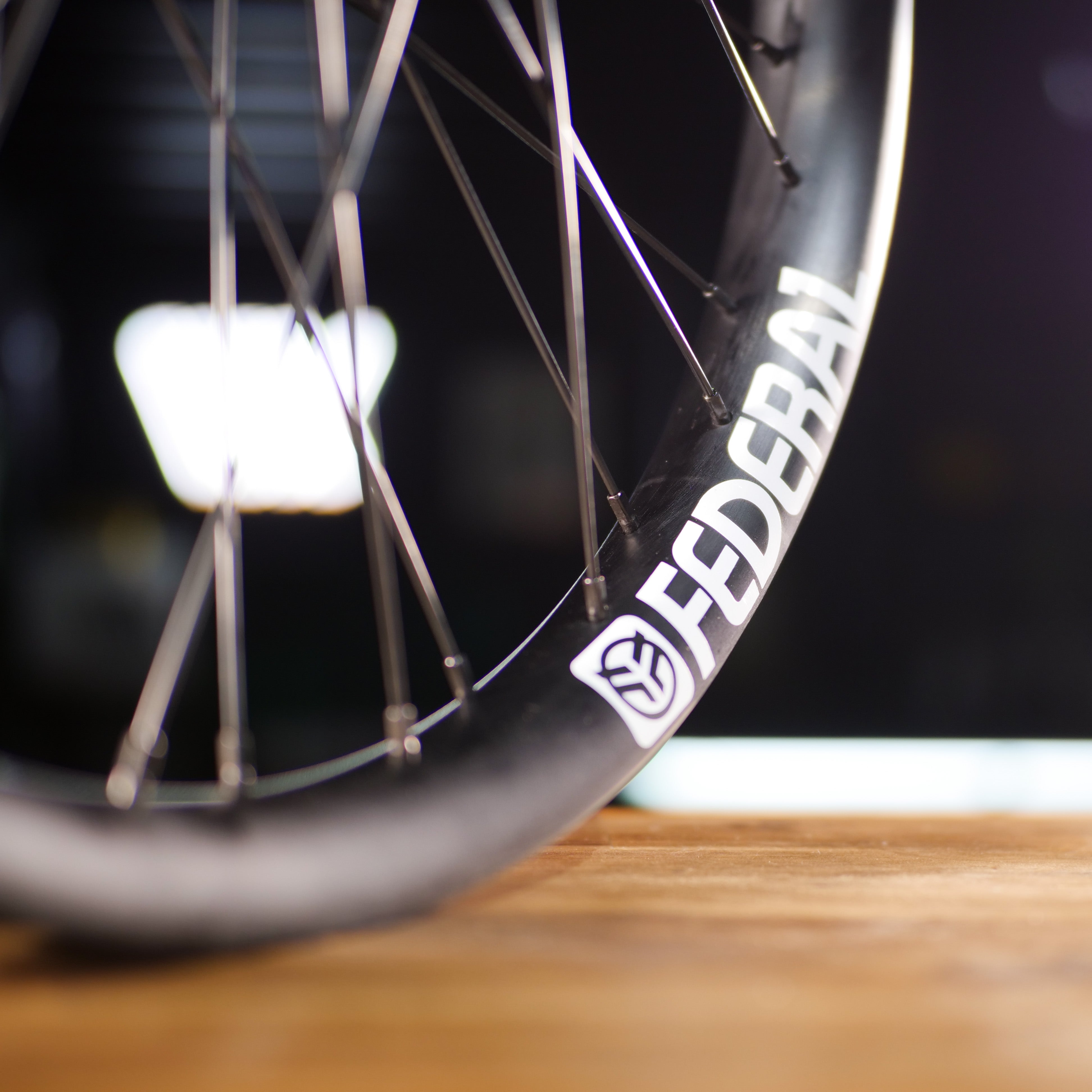 Close-up of the Federal X Eclat FM Shift Hybrid Custom Wheel with a black "FEDERAL" rim and shield logo, metal spokes, and hybrid hub, displayed on a wooden surface.