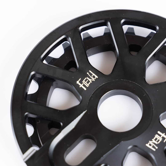 Close-up of a Fiend Omicron Guard Sprocket with a series of cutouts and geometric patterns, crafted from 6061 aluminium. It features a central hole and several surrounding holes, with a partially visible logo in white text.