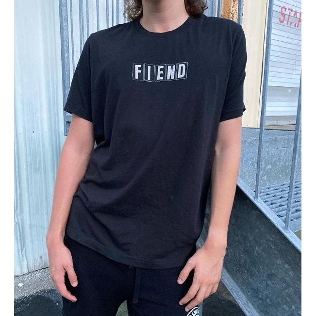 A person in a black Fiend Ransom Vintage Shirt, made from 100% airlume cotton with "FIEND" stylishly discharged printed on it, poses confidently before a metal fence.
