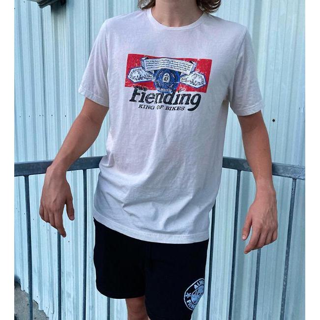 A person confidently stands before a corrugated metal wall, wearing a unisex Fiend Shirt King Of Bikes in vintage white with soft touch printing and black shorts, offering style and comfort for everyone.