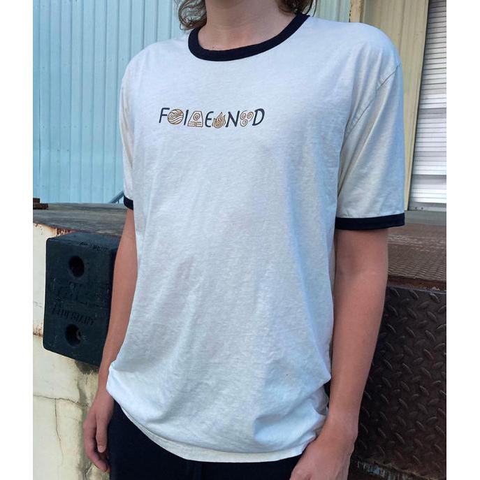 A person wears the Fiend Shirt Raekes DT Natural— a ringer-style t-shirt with a white body and black trim, featuring abstract text. The setting is an industrial backdrop with a metal wall, adding an edgy vibe typical of rider-owned brands.