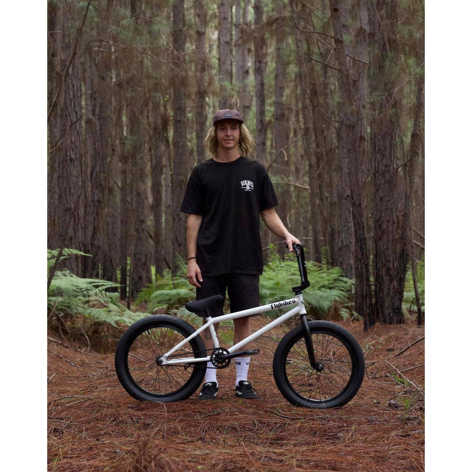 In a wooded area, someone wears a dark shirt, shorts, and a cap while holding a white bike with black wheels made of multi-butted 4130 CrMo. The sturdy frame features Fly Bikes Dove Bars and includes a Lifetime Warranty for added peace of mind.