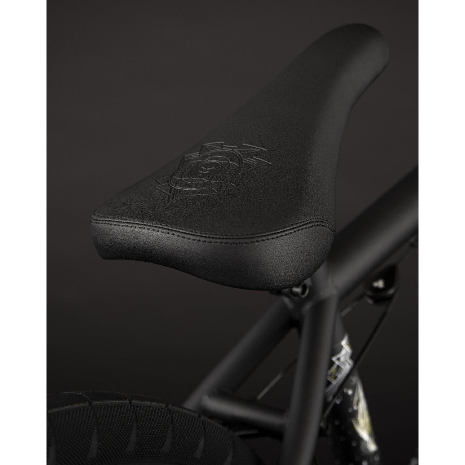 A close-up of the Fly Bikes Electron 20 Inch Bike features a black bicycle seat with an embossed design, mounted on a Cr-Mo frame, set against a dark background.