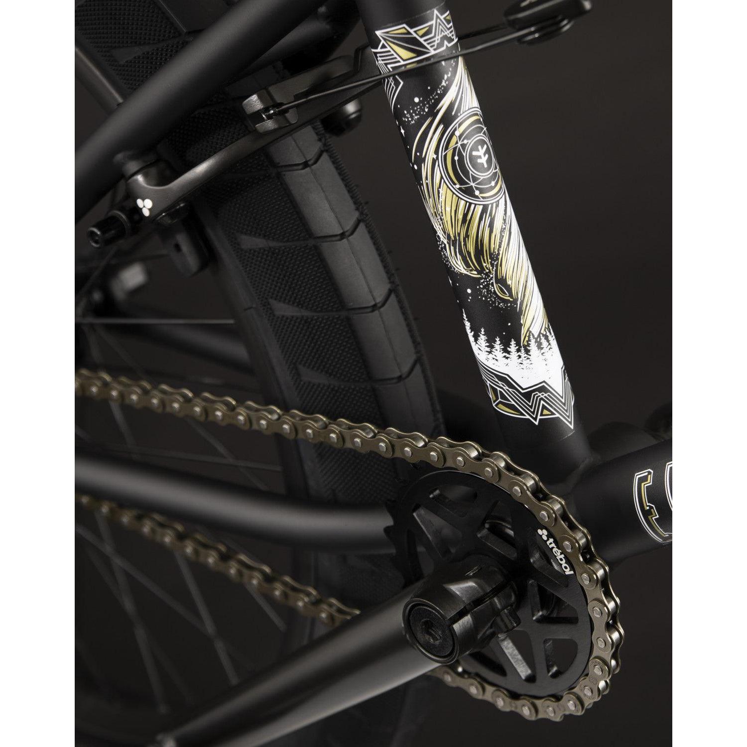 A close-up of the Fly Bikes Electron 20 Inch Bike highlighting its decorated frame made from Cr-Mo material, equipped with Trebol Tires, a shimmering gold chain, and intricate graphics featuring a nature-themed design.
