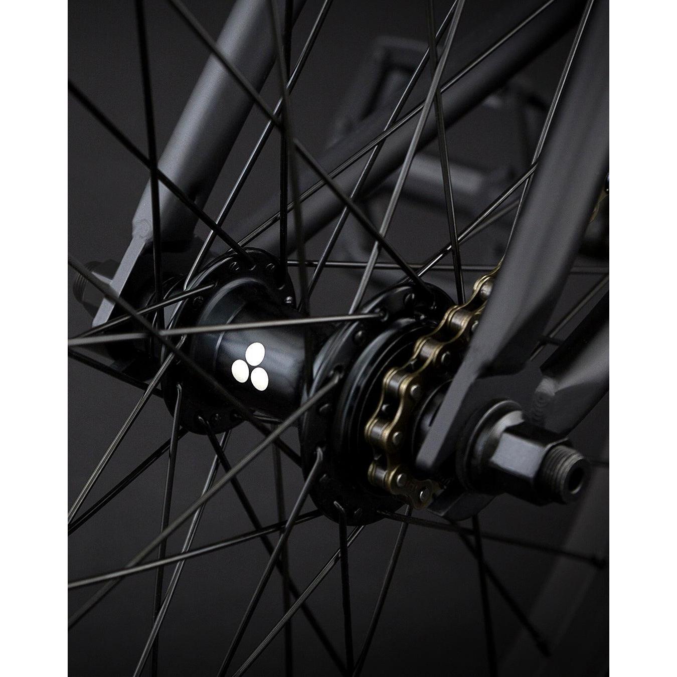 A close-up of the Fly Bikes Neutron 20 Inch Bike's rear wheel hub features a logo, highlighting its dark spokes and chainset against a dark background. The CrMo frame enhances durability and strength, contributing to the bike's sleek design.