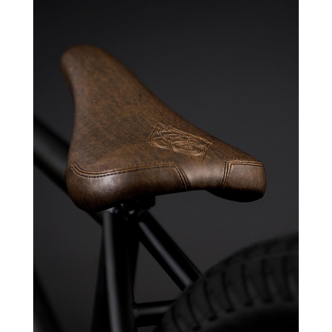 Close-up of a brown seat, engraved with the Fly Bikes logo, mounted on the sleek CrMo frame of the Neutron 20 Inch BMX bike, set against a dark background.