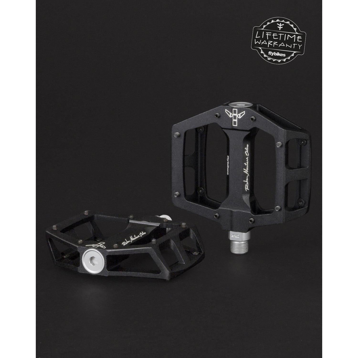 Two black Fly Bikes Ruben Aluminum Pedals with silver detailing are set against a dark backdrop. One is upright, highlighting its CNC Chromoly axle. The other is flat. A "Lifetime Warranty" logo in the top right promises durability with precision bearings.