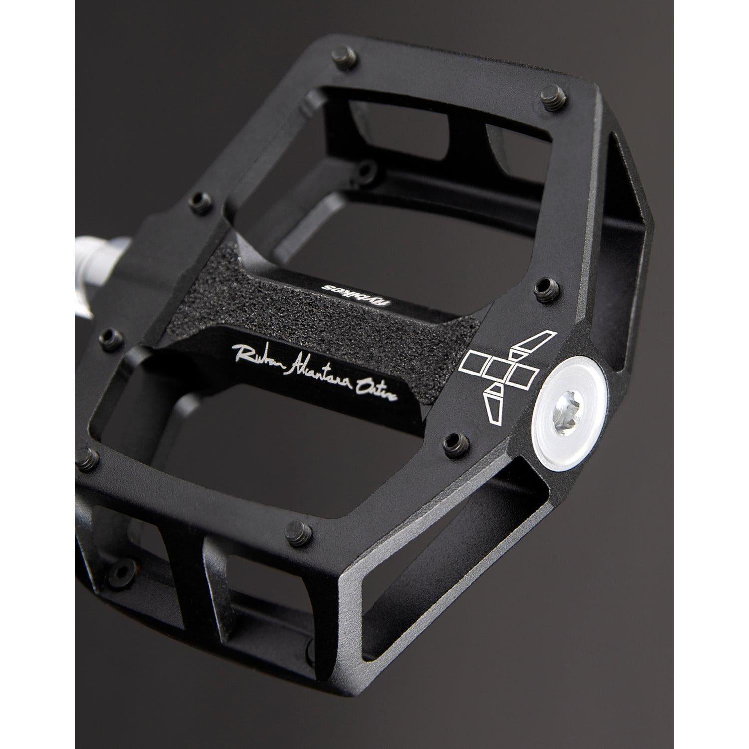 Close-up of a sleek black Fly Bikes Ruben Aluminum pedal with a metallic body and CNC Chromoly axle, featuring precision bearings. The visible branding stands out against the dark background.