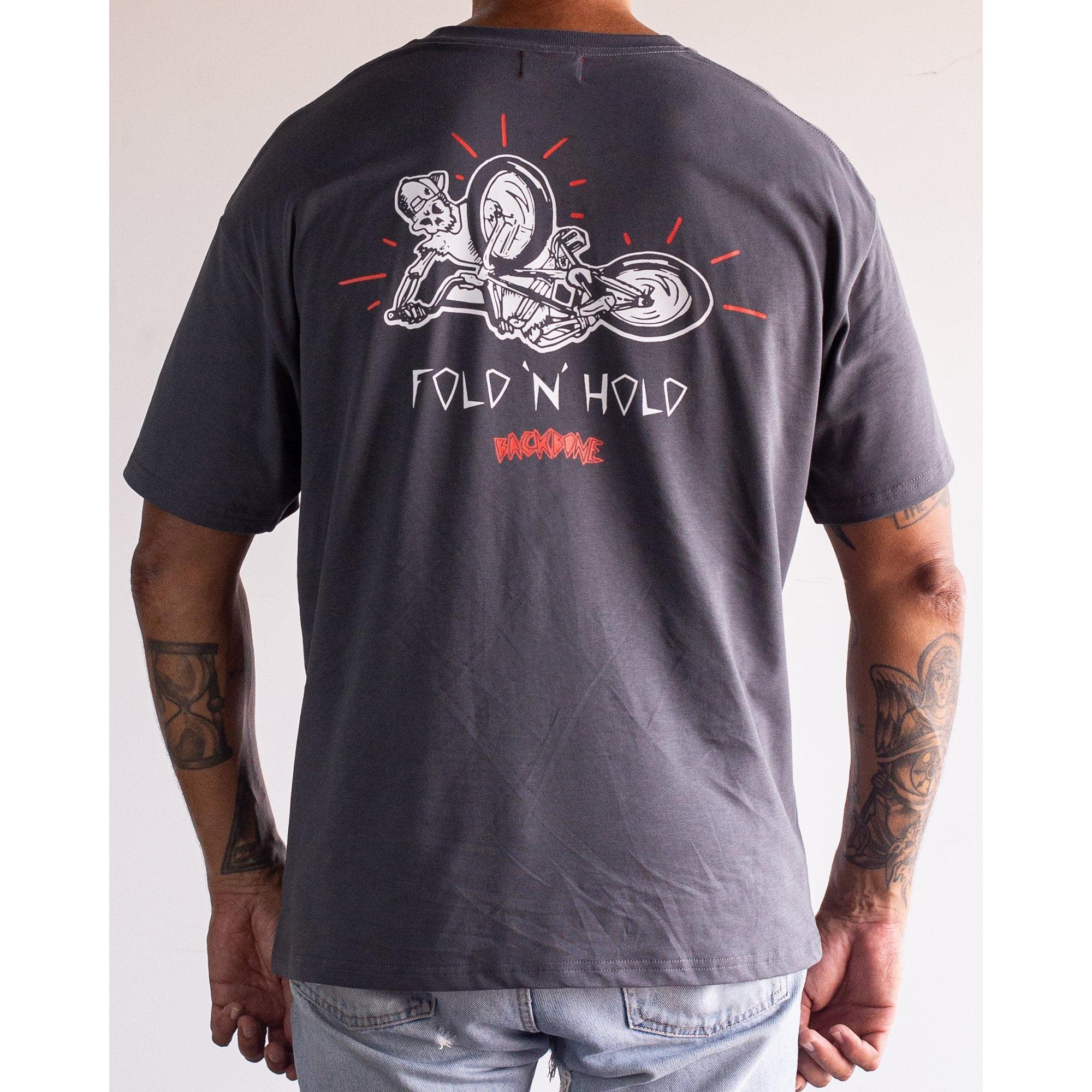 Person wearing the Backbone Fold 'N' Hold t-Shirt in dark gray, made from premium cotton, with a motorcyclist stunt graphic and “FOLD N’ HOLD” plus “BACKBONE” text on the back.
