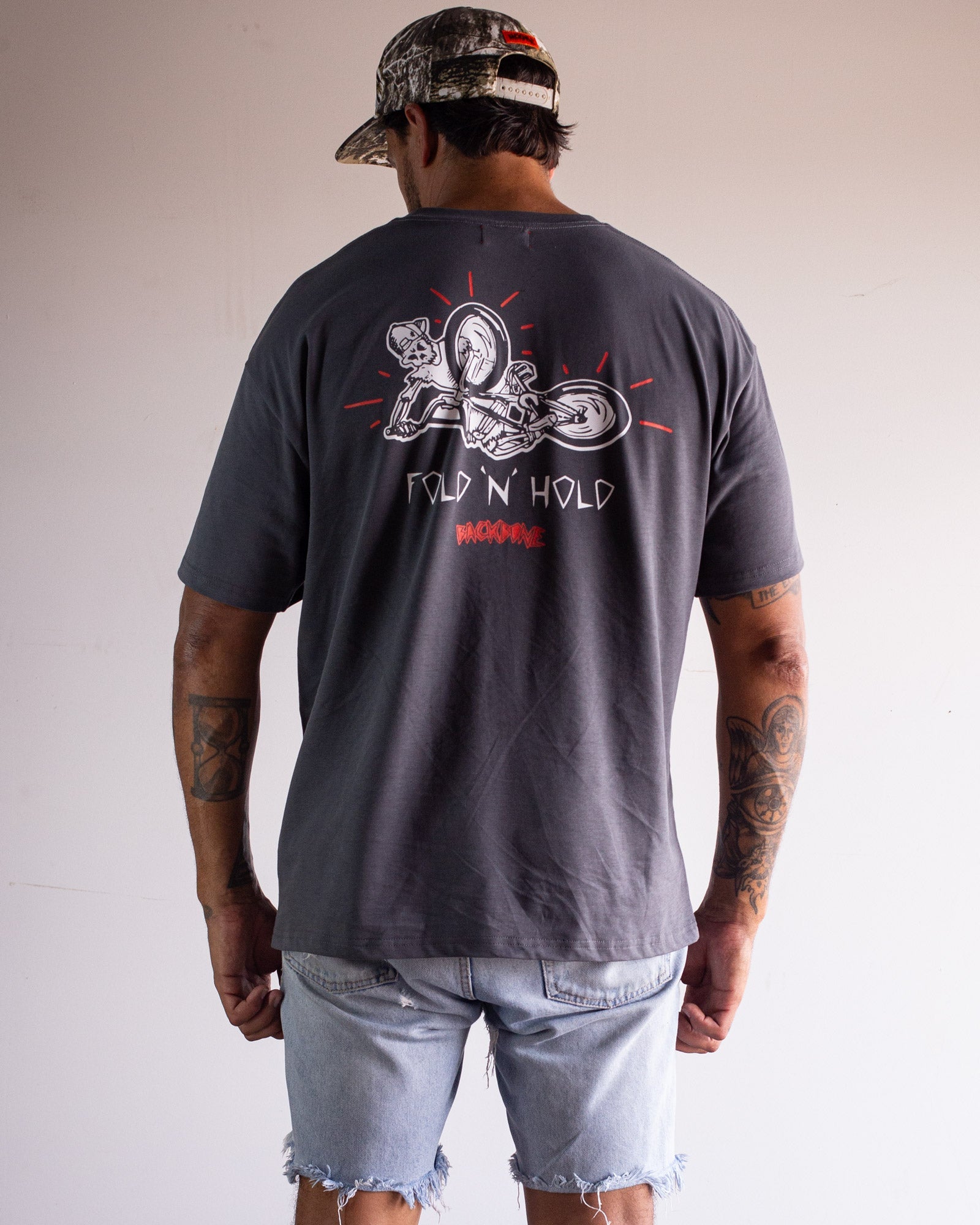 A man in a camo cap, light denim shorts, and a dark Backbone Fold 'N' Hold t-Shirt with a motorcycle graphic stands with his back to the camera.