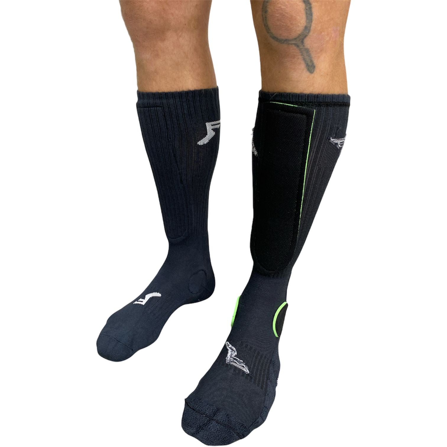 A person wearing black socks—on the right foot, an FP Painkiller Socks Crew Cut (Pair) style sock with a black shin guard and green trim; on the left, a plain black sock.