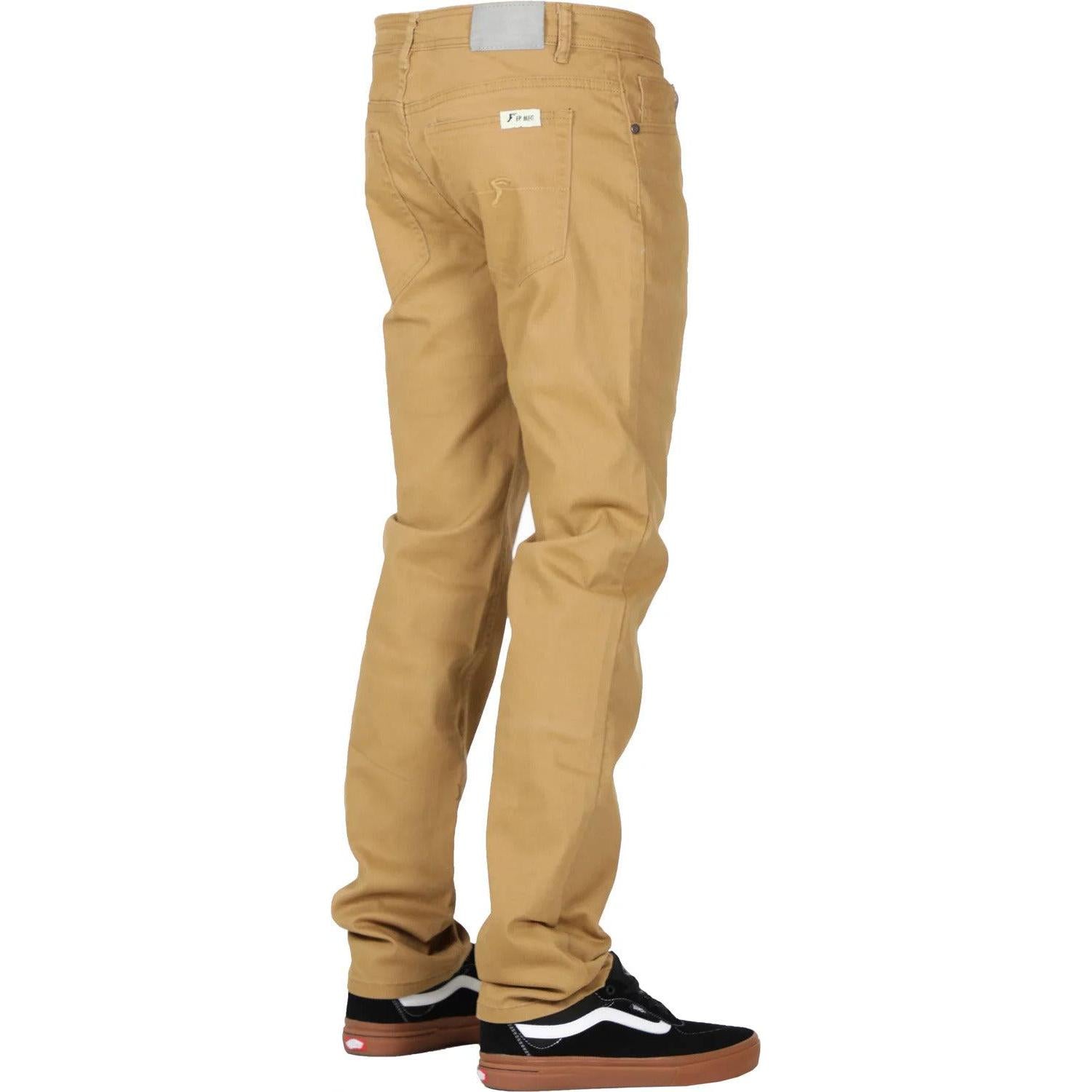 A person is standing, wearing khaki **FP Pants Relaxed Fit Chino Bone** and black sneakers with white accents. The pants, made of stretch fabric, have a small white label near the back pocket. The background is white.
