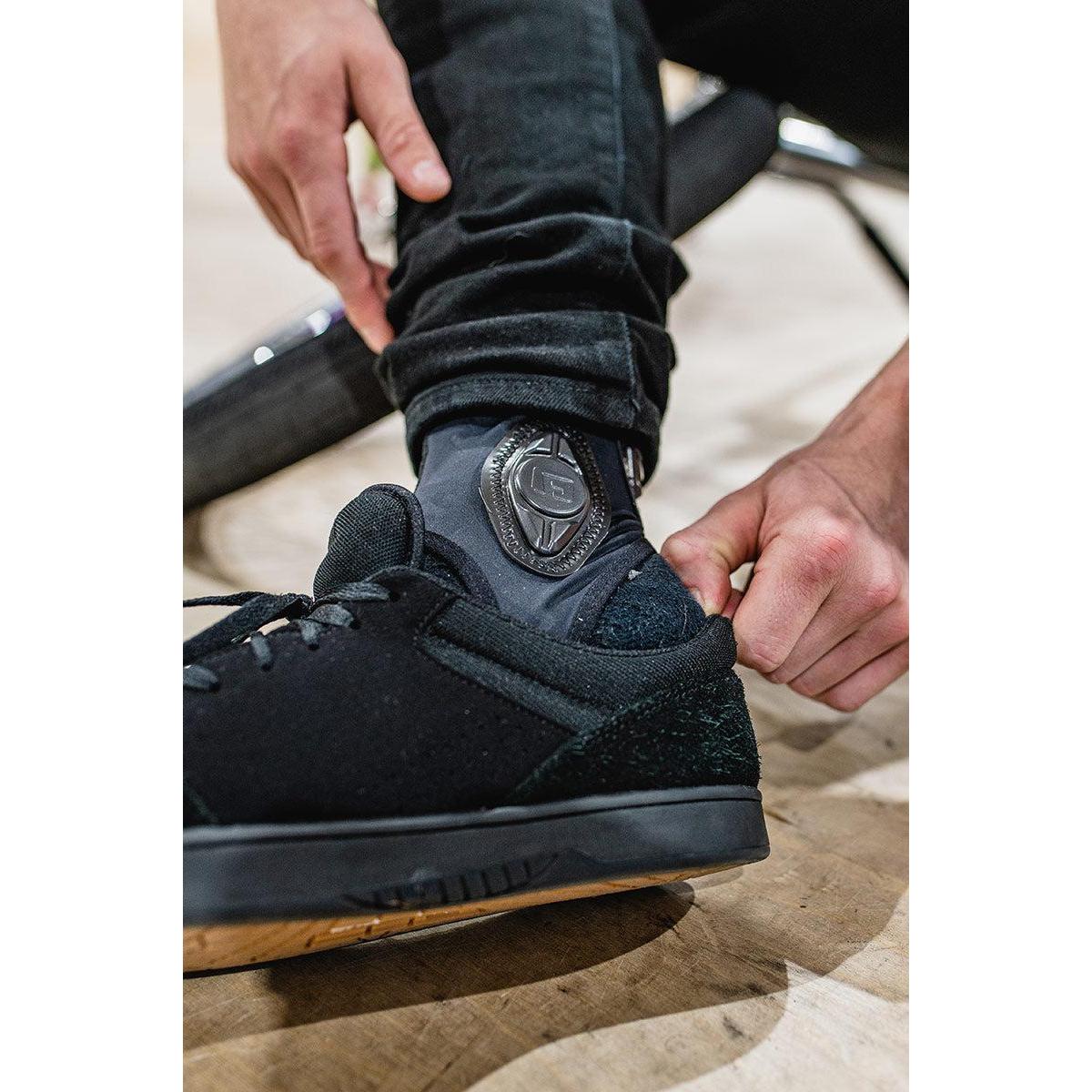 A person adjusts their G-Form Pro-X Ankle Guards, featuring SmartFlex pads for enhanced impact protection, while wearing matching black socks and shoes.