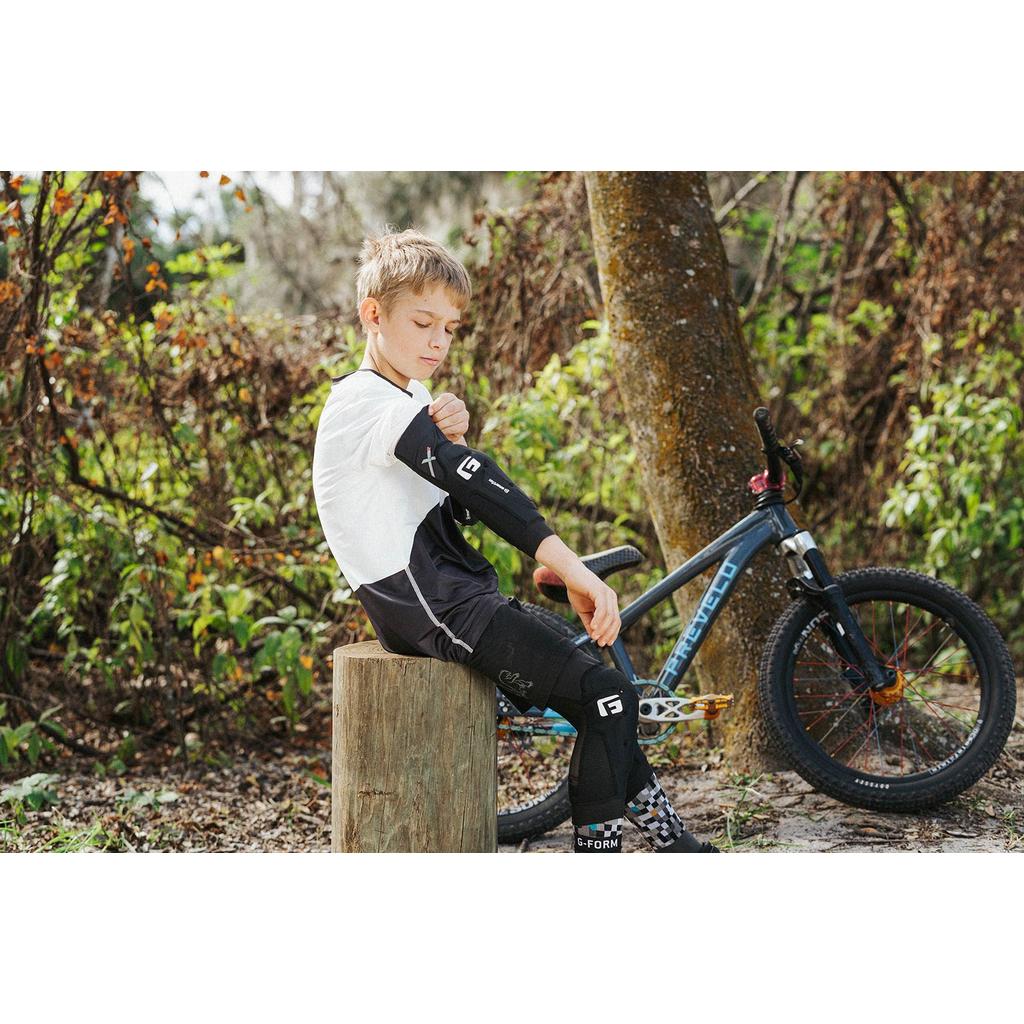 A boy sits on a tree stump in a wooded area, adjusting his G-Form X4 Elbow Guard, with a mountain bike resting nearby.