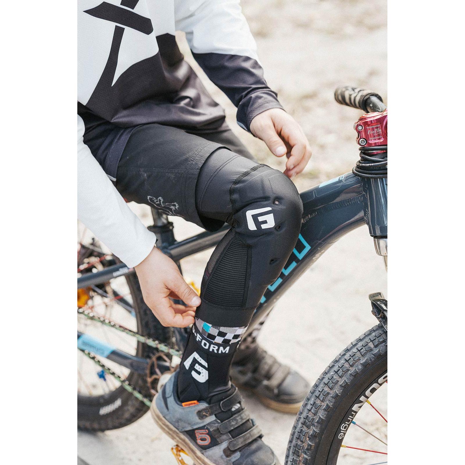 Wearing G-Form X4 Knee Guards with SmartFlex padding and cycling gear, a person sits on a black mountain bike and adjusts their checkered socks.