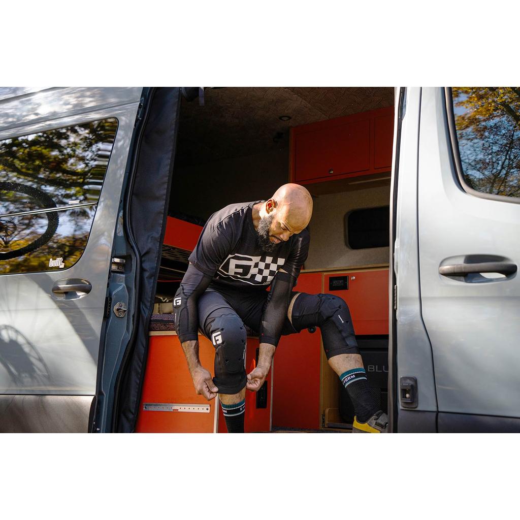 Wearing G-Form X4 Knee Guards, a person adjusts their gear while seated at the entrance of a camper van with orange cabinets visible inside.