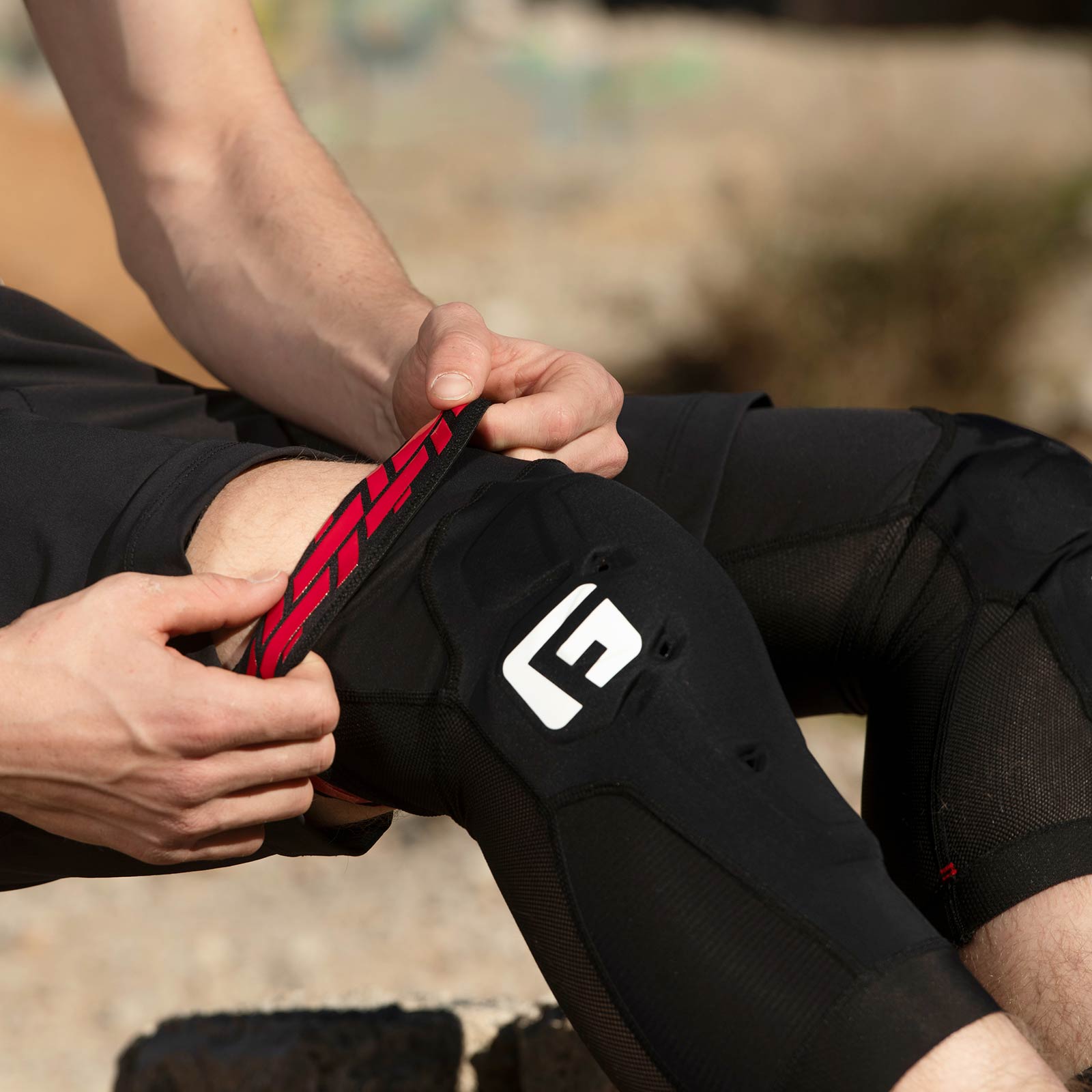 A person adjusts the strap of a G-Form X4 Knee Guard, CE-1621-1 certified and featuring a white logo, while wearing black athletic shorts outdoors.