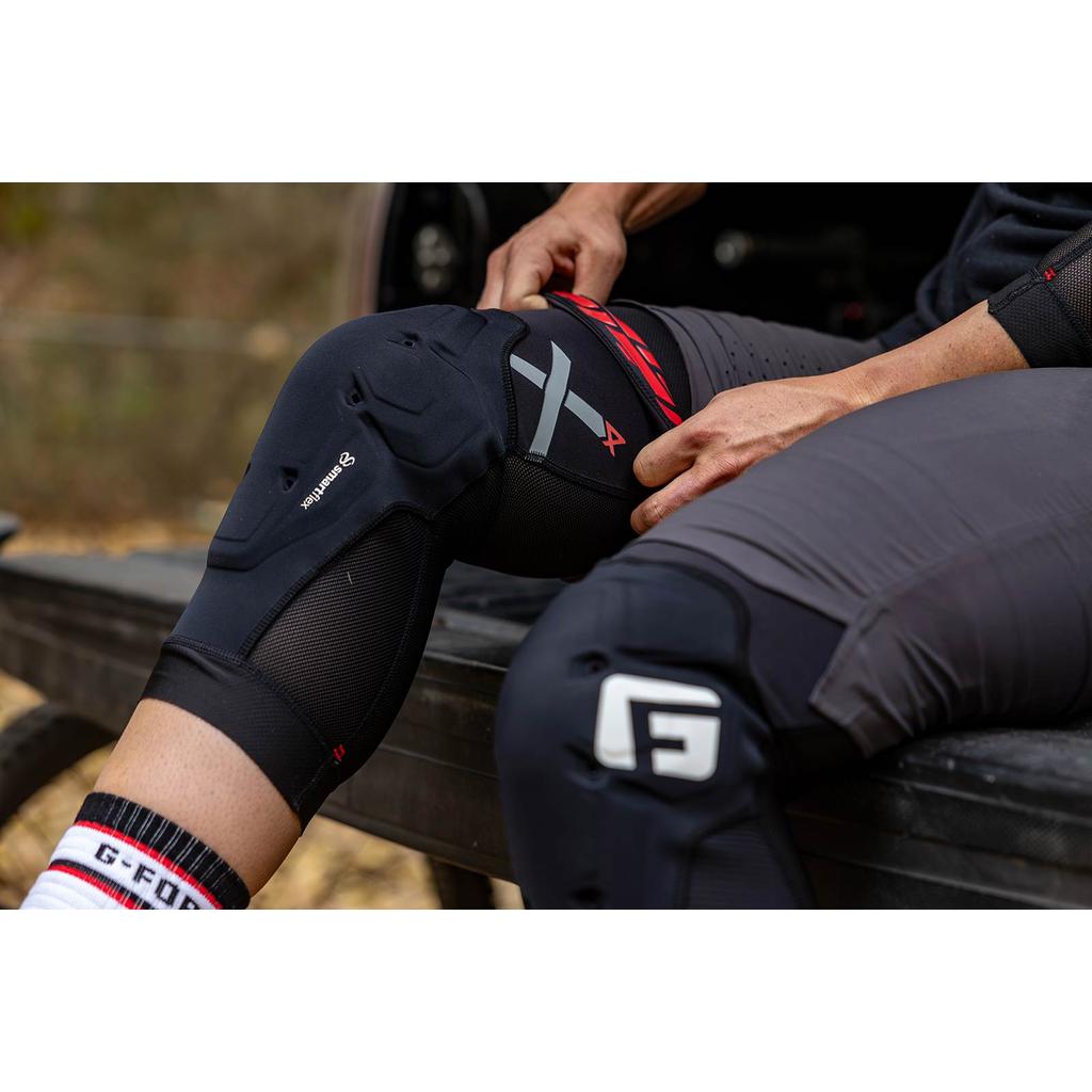 Person adjusting black G-Form X4 Knee Guard while sitting on a vehicle edge, wearing dark pants and white socks with “G-FORM” logo. These CE-1621-1 certified knee guards deliver top-level protection and comfort.