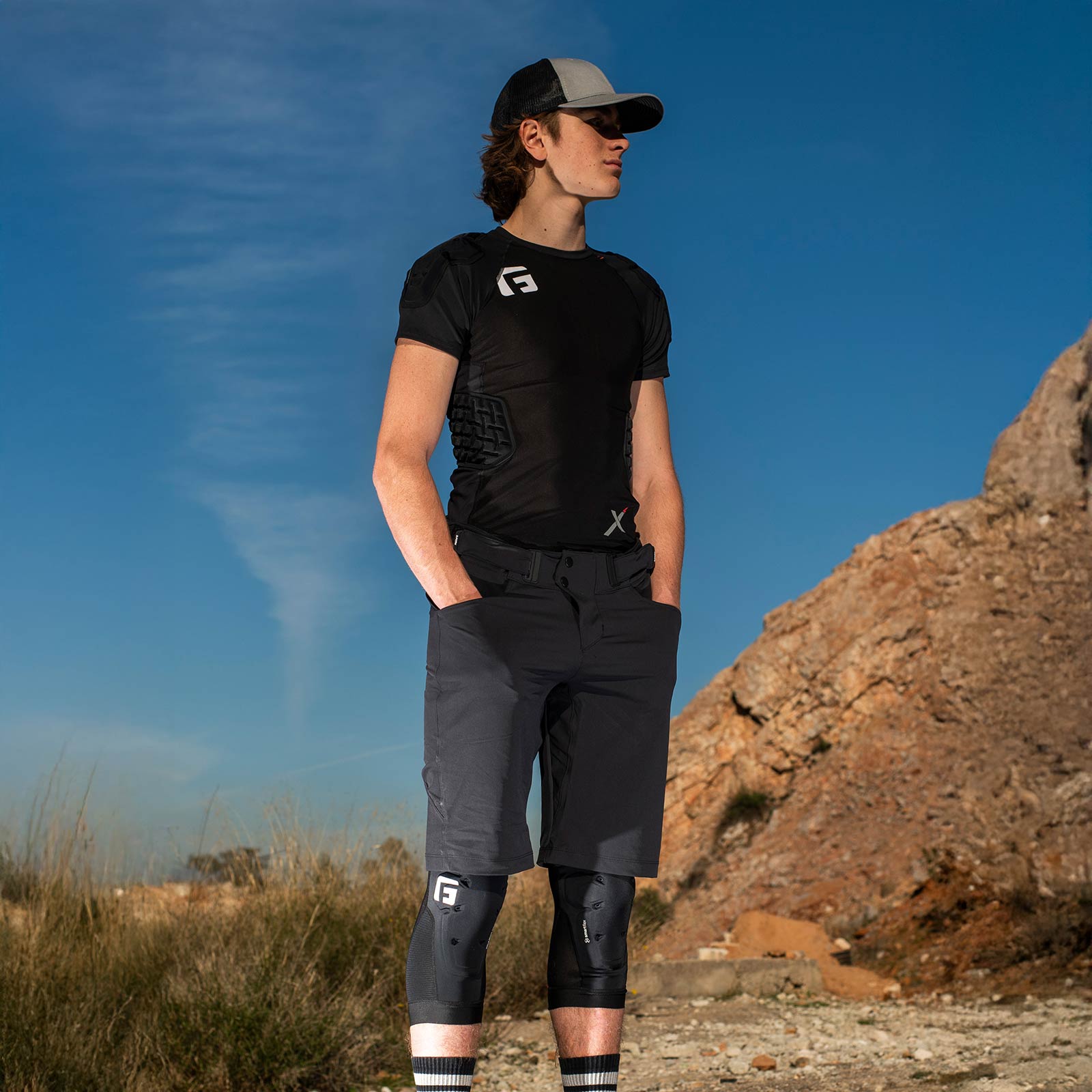 Wearing a G-Form X4 Padded Shirt, knee pads, and a cap, the person stands outdoors on rocky ground beneath a clear blue sky.