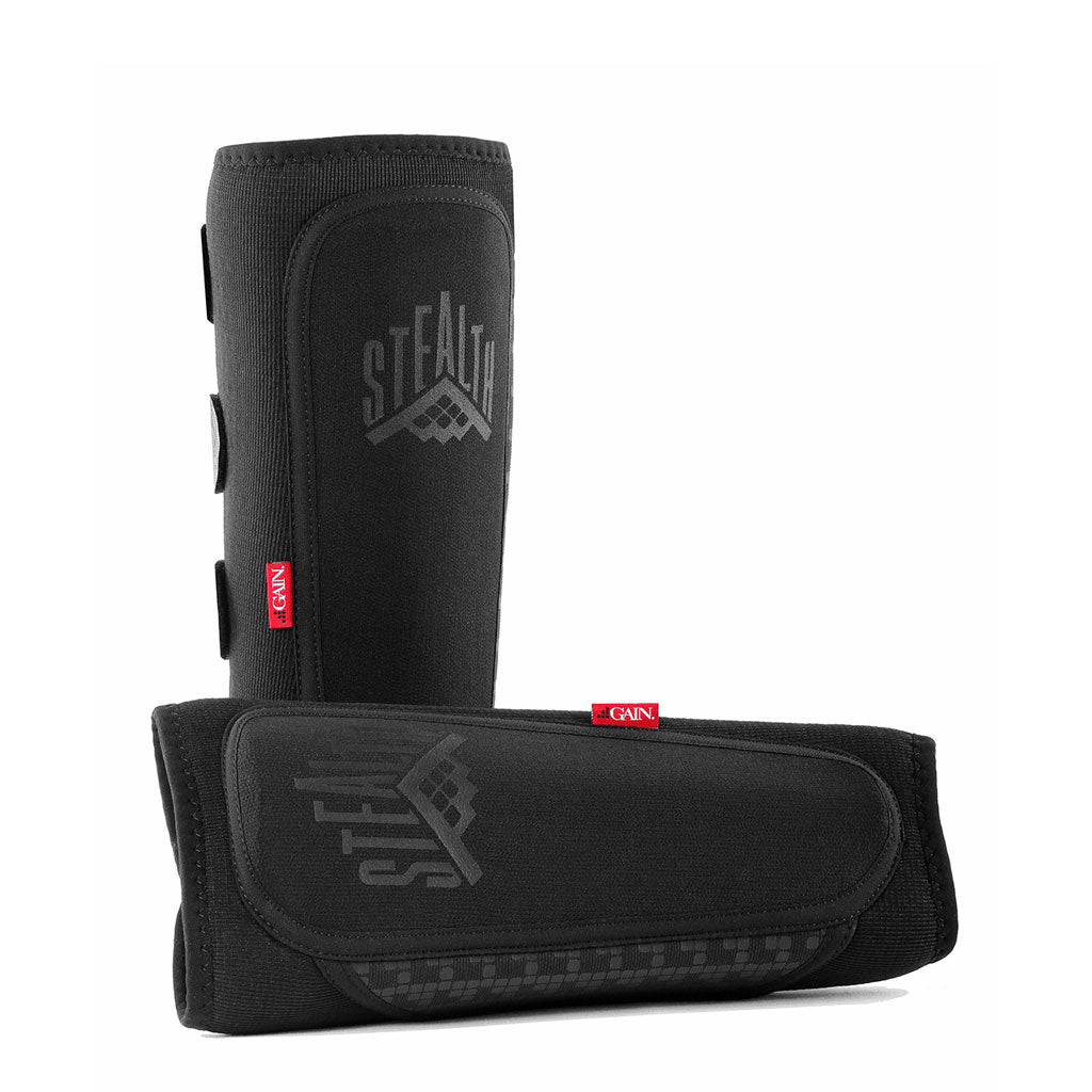 A pair of black GAIN STEALTH Shin Guards with a geometric "STEALTH" logo, made from duraTec neoprene, feature velcro straps and red CANI tags, displayed upright and horizontally on a white background.