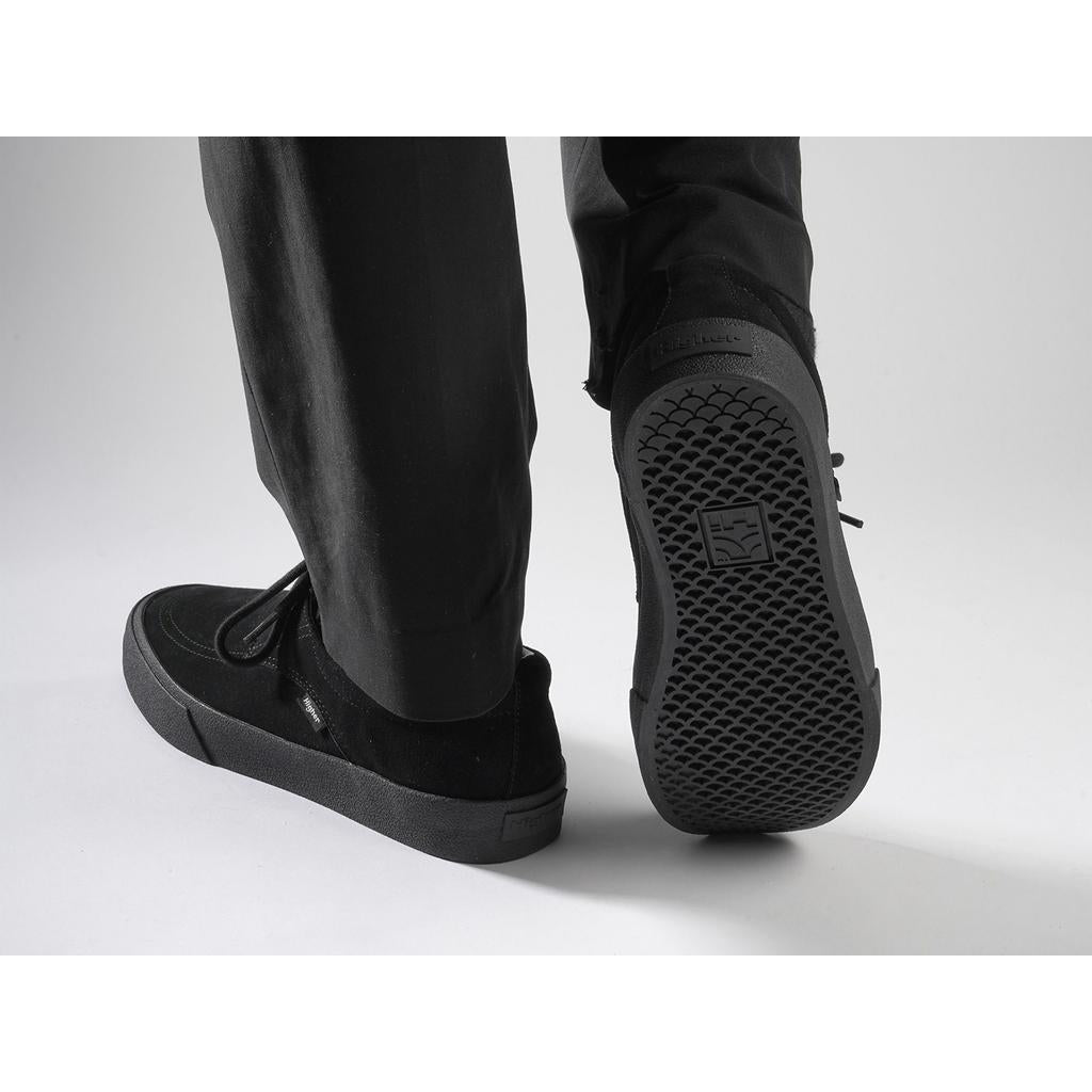 A person wearing Higher Salem Suede Slip On Shoes in black, seen from behind; the visible shoe sole displays a textured pattern and logo, and the shoes feature a reinforced toe box.