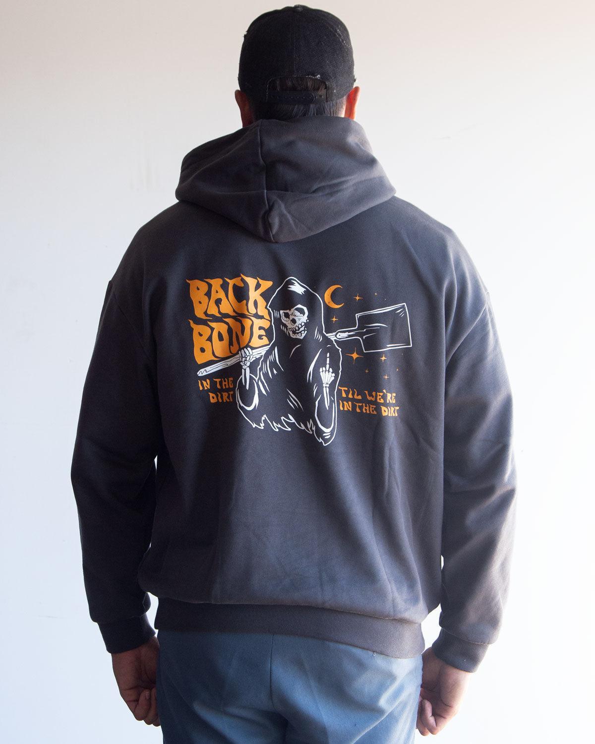 A person wearing the Backbone In the Dirt Hoodie—a heavyweight, oversized hoodie featuring a graphic of a skeleton and moon, with the text “BACK BONE IN THE DIRT ALL WEEK IN THE DIRT” printed on the back.