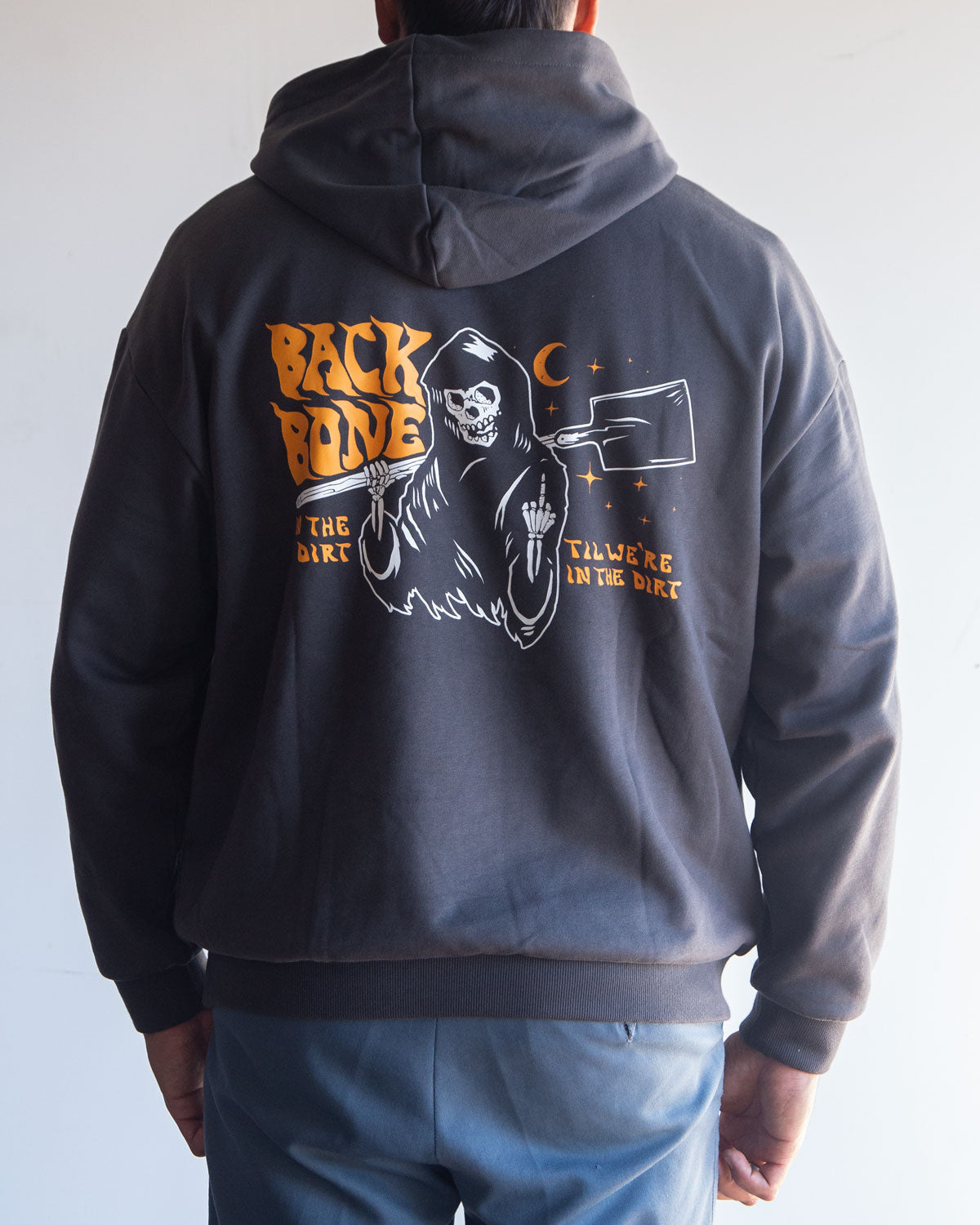 A person is wearing the Backbone In the Dirt Hoodie, an oversized black hoodie featuring a Grim Reaper graphic holding a cleaver, with “BACKBONE” and “I’ll Work In The Dirt” text on the back.