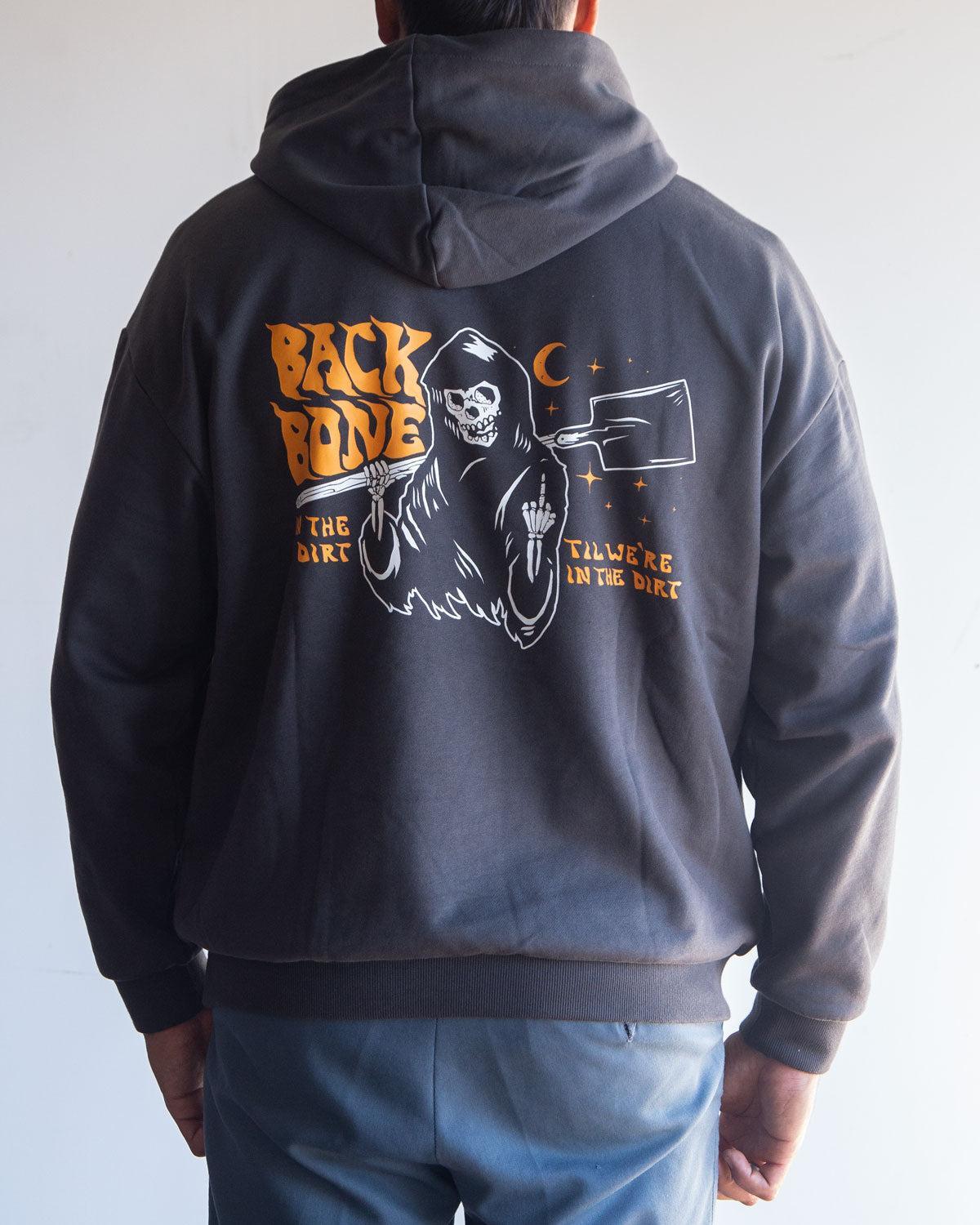 A person is wearing the Backbone In the Dirt Hoodie, an oversized black hoodie featuring a Grim Reaper graphic holding a cleaver, with “BACKBONE” and “I’ll Work In The Dirt” text on the back.