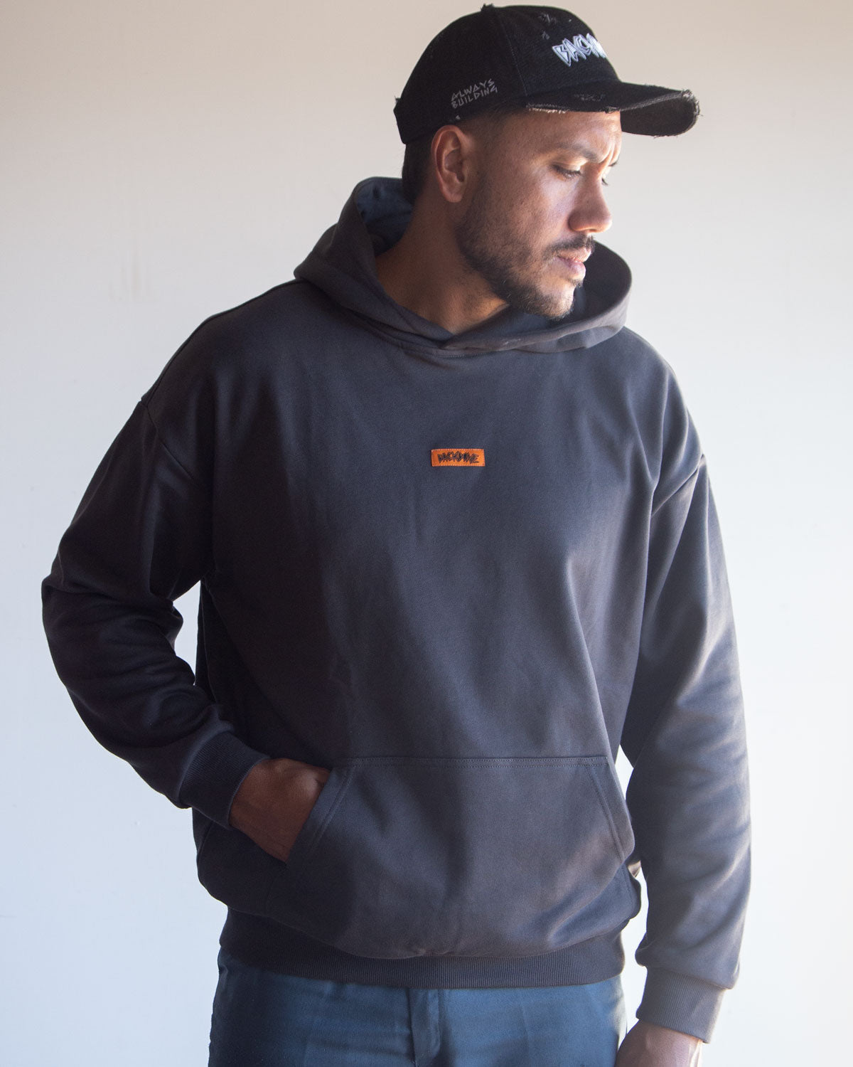 A man wearing the Backbone In the Dirt Hoodie—a black heavyweight hoodie with an orange patch—paired with a black cap, stands with one hand in his pocket, turned slightly to the side.