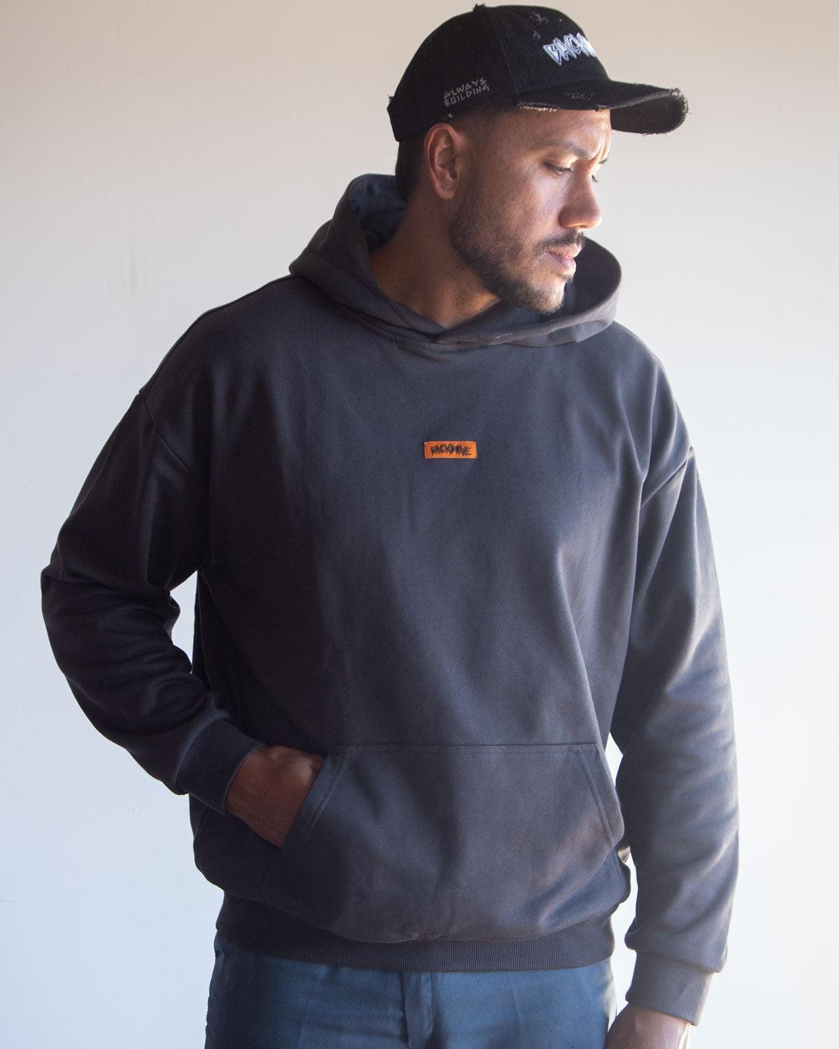 A man wearing the Backbone In the Dirt Hoodie—a black heavyweight hoodie with an orange patch—paired with a black cap, stands with one hand in his pocket, turned slightly to the side.
