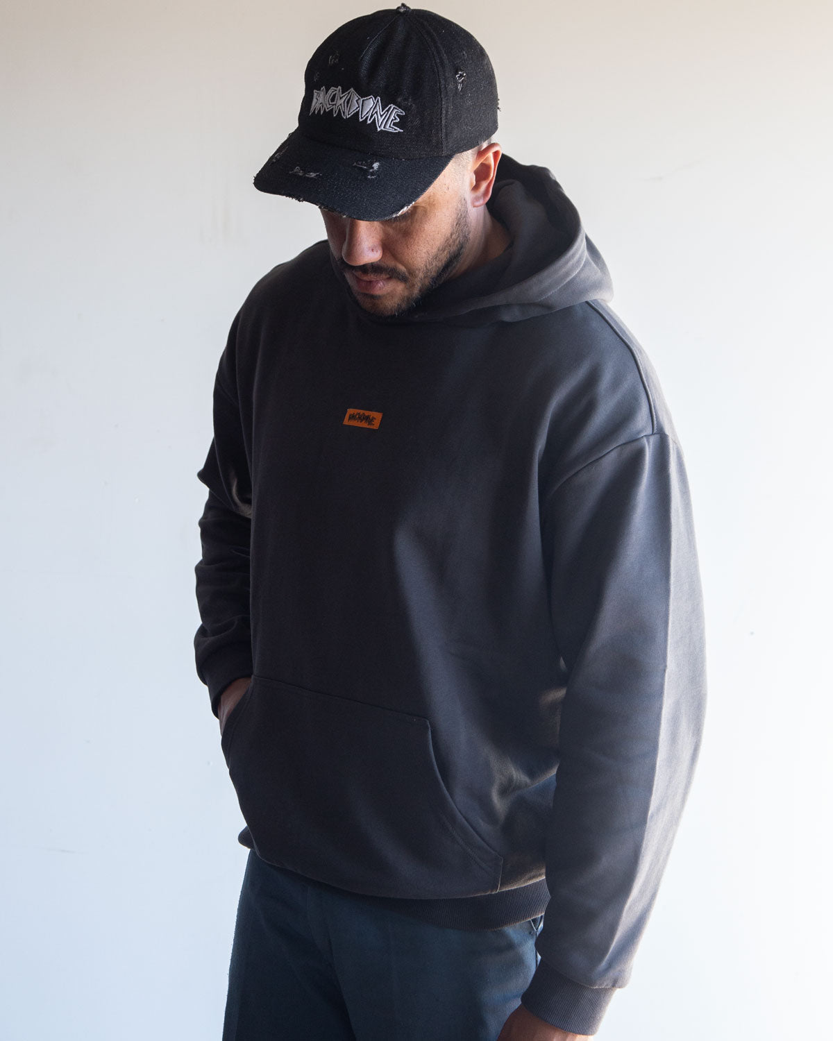 A man wearing the Backbone In the Dirt Hoodie—a black, heavyweight style with a small orange label—paired with a black cap featuring white text, stands against a plain light background looking down.
