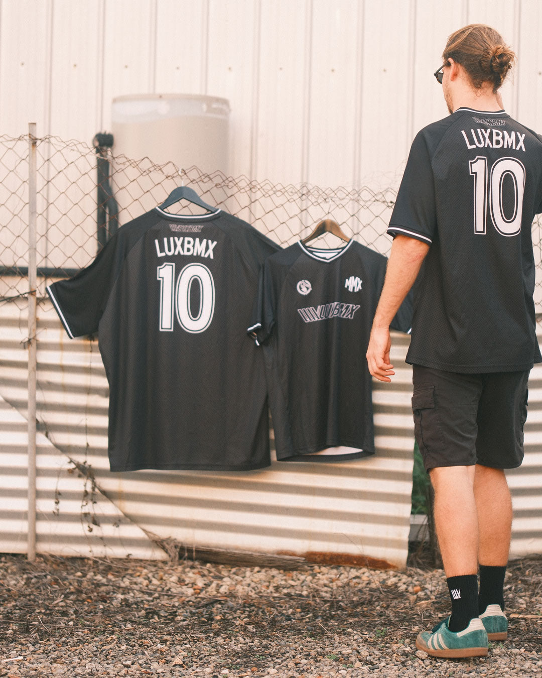 A person in a black LUXBMX MMX Soccer Jersey - Custom Back Print stands outdoors near two identical jerseys hanging on a metal fence.