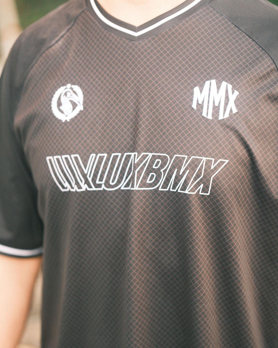 A person wears the LUXBMX MMX Soccer Jersey - Custom Back Print, featuring black, breathable fabric, "LUXLUXBMX" white text, "MMX" abbreviations, and a circular chest logo.