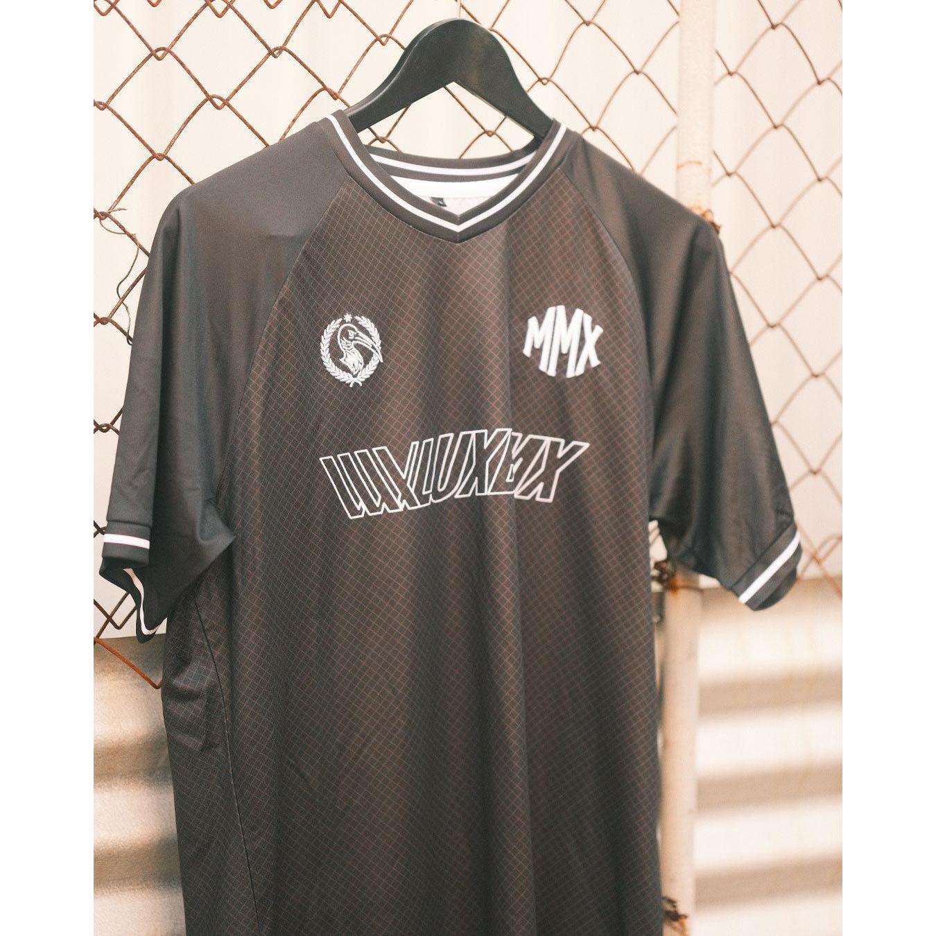 The LUXBMX MMX Soccer Jersey - Custom Back Print features a black and gray design with white trim and logos, displayed on a black hanger against a chain-link fence—perfect for those seeking a custom BMX jersey style.