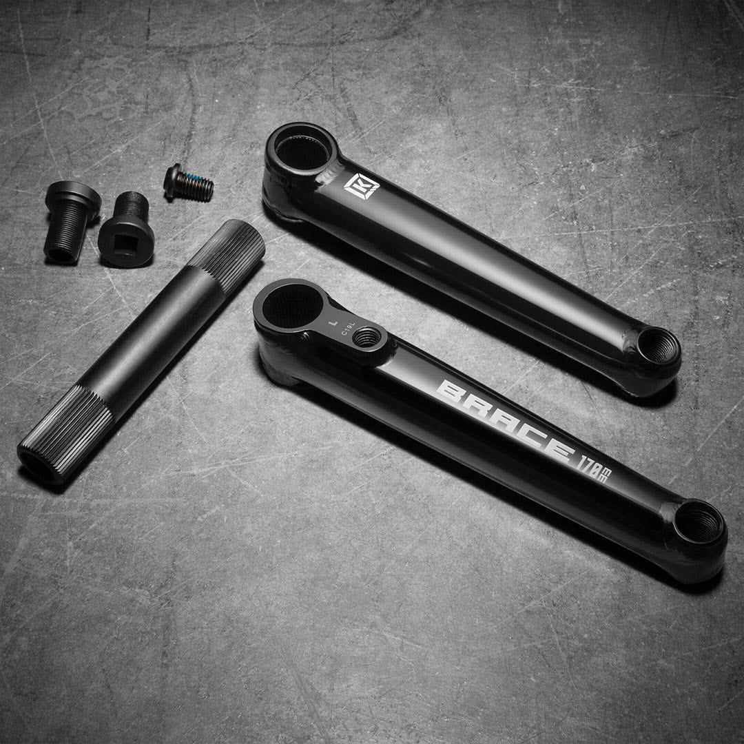 On a gray surface, the Kink Brace Cranks are displayed, showcasing two black crank arms marked "BRACE 178A" with a 3-piece design, a 22mm hollow bottom bracket spindle, and small parts such as bolts and caps.