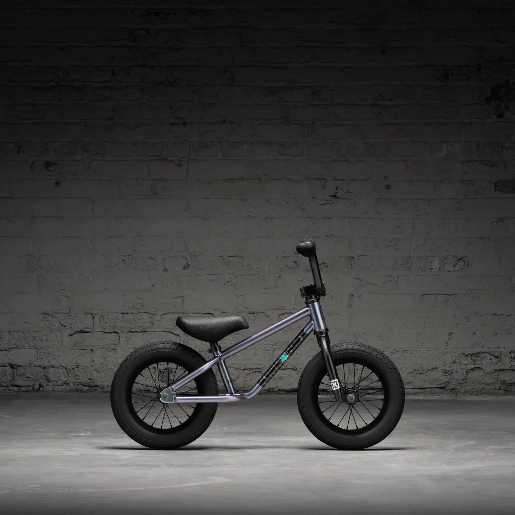 A Kink Coast 12 Inch Balance Bike (2026) with a purple frame and black seat, tires, and handlebars is positioned against a textured gray brick wall.