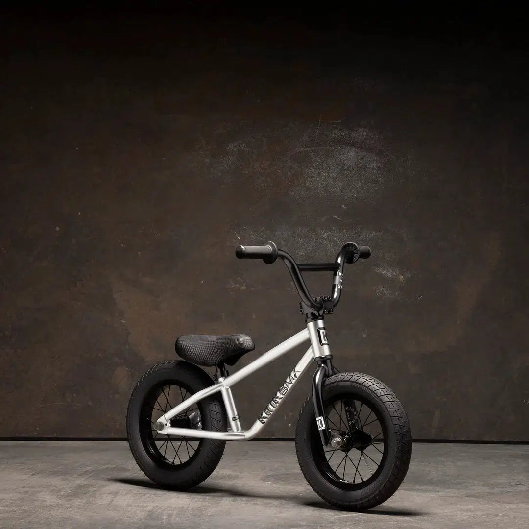The Kink Coast 12 Inch Balance Bike, featuring a small silver frame with black handlebars, seat, and tires, stands out against a dark background.