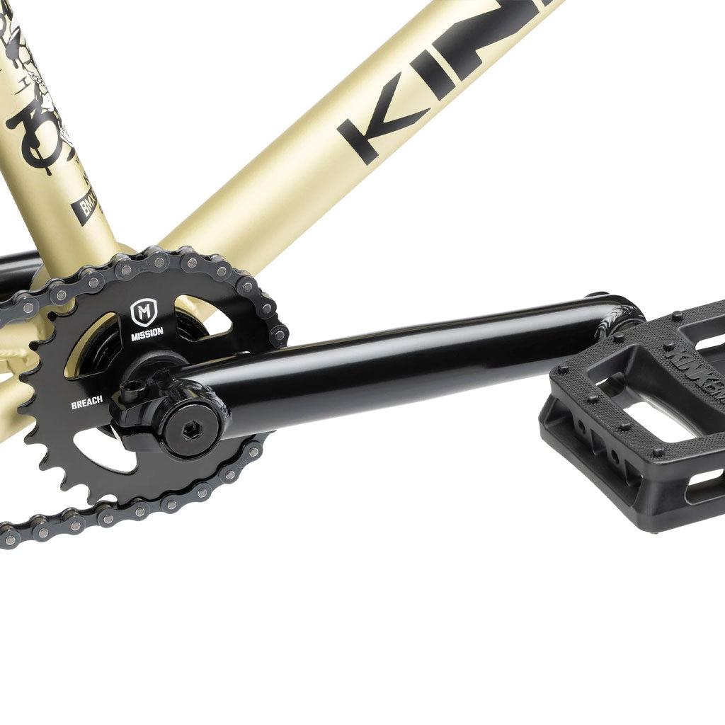 Close-up of the Kink Curb 20 Inch Bike's crankset and pedal, featuring a chainring labeled "Mission" and "Reach," attached to a beige and black frame emblazoned with the "Kink" branding, highlighting its high-quality BMX components.